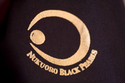 Gold emblem on black cloth, saying Nukuoro Black Pearls, with a logo shaped like a crescent with a pearl in between the two pincers