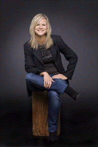 picture of Johnson Mandell, a woman, in a dark blazer, dark tee, jeans, sitting on a tree trunk